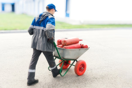 Man in work clothes carries red fire extinguishers in wheelbarrow. Industrial recycling and recharging of fire extinguishers. Fire extinguishing agent.の写真素材