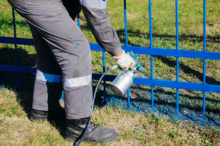 Worker in overalls paints external metal fence from spray gun on summer day. Kind of spray gun in hand. Industrial painting of fence. Professional painter at work. Authentic workflow..の写真素材