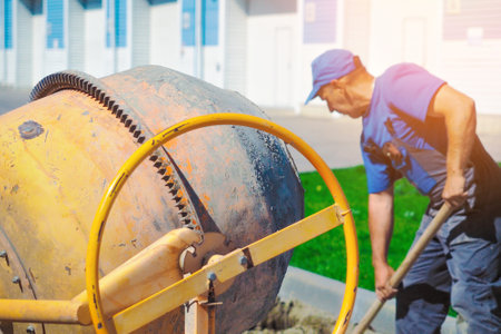 Uniformed construction worker works on construction site on summer day. Elderly bricklayer shovels cement and sand into concrete mixer. Authentic workflow..の写真素材