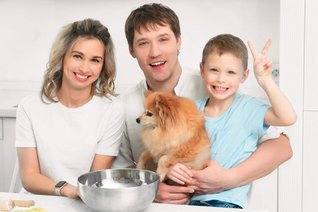Cheerful happy family of three together with Spitz dog sit at kitchen table and laugh. Portrait of young Caucasian family. Son sits on his fathers lap and smiles..の写真素材