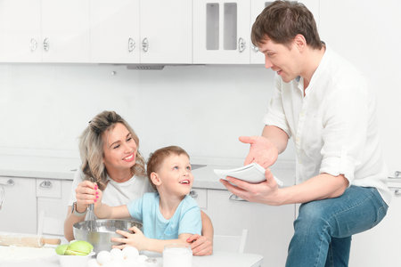 Young Caucasian family of three cooks lunch or festive cake in kitchen according to recipe. Happy family at table in bright kitchen..の写真素材