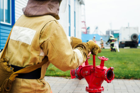Fire rescuer in helmet and protective clothing sits next to fire hydrant outside on street. View...