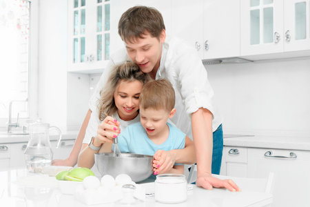 Young Caucasian family of three cooks lunch or festive cake in kitchen according to recipe. Happy family at table in bright kitchen..の写真素材