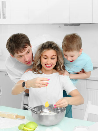 Cheerful family in kitchen breaks egg into plate. Parents with young son together prepare delicious dessert..の写真素材