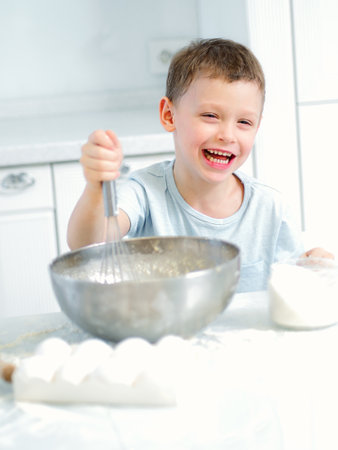 Child cooks food. Caucasian boy sits at table in kitchen and kneads dough in cup with hand whisk. Cheerful and positive kid cooks pie and laughs. Young cook or chef. Portrait of child..の写真素材