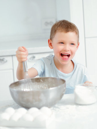 Child cooks food. Caucasian boy sits at table in kitchen and kneads dough in cup with hand whisk. Cheerful and positive kid cooks pie and laughs. Young cook or chef. Portrait of child..の写真素材