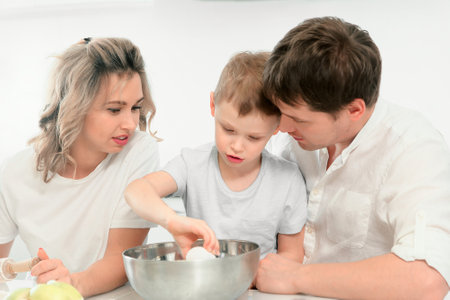 Young Caucasian family of three cooks lunch or festive cake in kitchen according to recipe. Happy family at table in bright kitchen..の写真素材