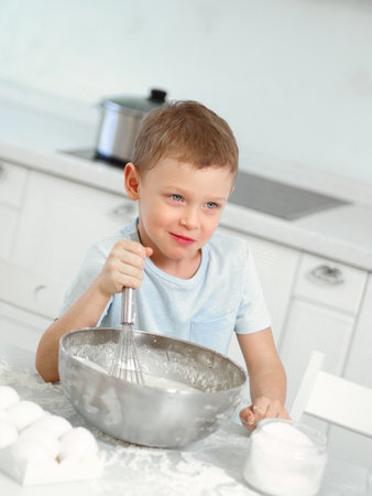 Child cooks food. Caucasian boy sits at table in kitchen and kneads dough in cup with hand whisk. Cheerful and positive kid cooks pie and laughs. Young cook or chef. Portrait of child..の写真素材