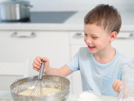 Child cooks food. Caucasian boy sits at table in kitchen and kneads dough in cup with hand whisk. Cheerful and positive kid cooks pie and laughs. Young cook or chef. Portrait of child..の写真素材