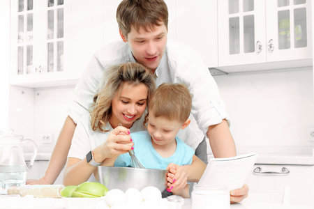 Young Caucasian family of three cooks lunch or festive cake in kitchen according to recipe. Happy family at table in bright kitchen..の写真素材