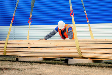 Slinger in helmet and signal vest unloads wooden boards on summer day outside on street. Construction material. Genuine workflowの写真素材