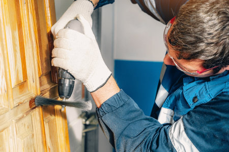 Man treats wooden surface. Worker in safety glasses grinds wooden door with metal brush. Professional repair of furniture and wooden products..の写真素材