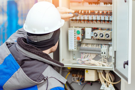 Professional electrician connects wires in electrical panel room outside. Worker in hard hat and winter clothes checks equipment with screwdriver. Back view.の写真素材