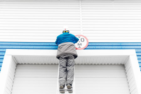 A male electrician stands on a ladder and replaces a light bulb in a street lamp on the gate of an industrial building or garage. Authentic workflow. View from the back from bottom to top.の写真素材