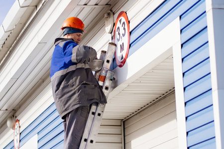 Male electrician stands on ladder and replaces light bulb in ceiling lamp on gate of industrial building or garage. Genuine workflowの写真素材