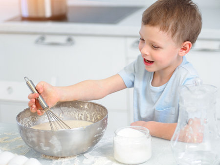 Child cooks food. Caucasian boy sits at table in kitchen and kneads dough in cup with hand whisk. Cheerful and positive kid cooks pie and laughs. Young cook or chef. Portrait of child..の写真素材