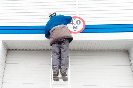 A male electrician stands on a ladder and replaces a light bulb in a street lamp on the gate of an industrial building or garage. Authentic workflow. View from the back from bottom to top.の写真素材