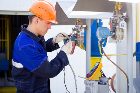Instrumentation and control engineer diagnoses the operation of gas distribution station equipment on a summer day. A professional worker in a hard hat and overalls diagnoses gas equipment sensors using a multimeter. Authentic worker portraitの写真素材