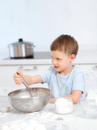 Cheerful boy plays chef at the table in the home kitchen. Caucasian child kneads the dough in a metal bowl. Playful kid in the kitchen.の写真素材