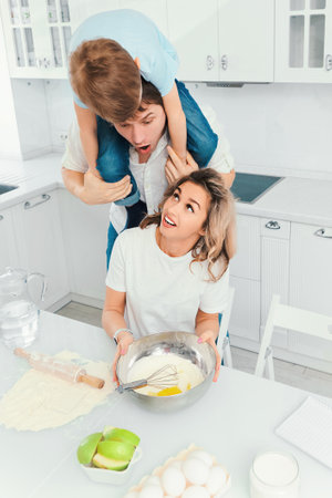 Young caucasian family mom dad and child are preparing birthday cake in home kitchen and having fun and fooling around. Cheerful parents together with their son start dough at kitchen table.の写真素材