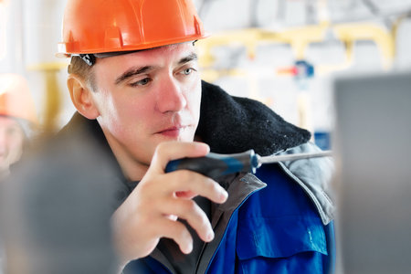 Close-up portrait of worker in hard hat with screwdriver during work process. Locksmith for repair and adjustment of industrial equipment and automation carefully makes diagnostics.の写真素材