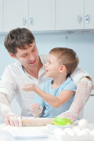 A young father and son are sitting at the kitchen table, covered in flour. A little boy and his father are making an apple pie together. A single father prepares a festive dessert with his little son.の写真素材