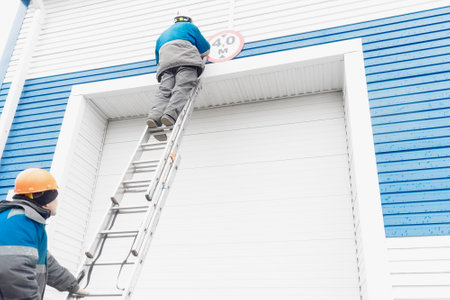 Two construction workers fix sign on automatic garage door for large vehicles. Worker stands on ladder and sets up height limit sign for entering garage.の写真素材