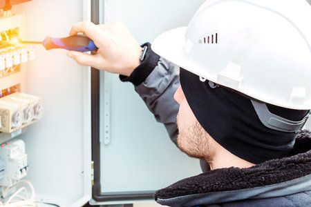 Professional electrician in hard hat connects wires with screwdriver in switchboard on winter day. Close-up portrait. Back view of head.の写真素材