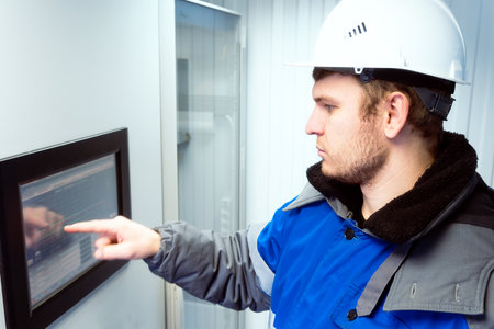 Engineer in hard hat and winter pea coat presses his finger on touch screen of modern production equipment indoors. Setting up automatic control system. Gas distribution station operator monitors gas flow readings. Photo. side view.の写真素材