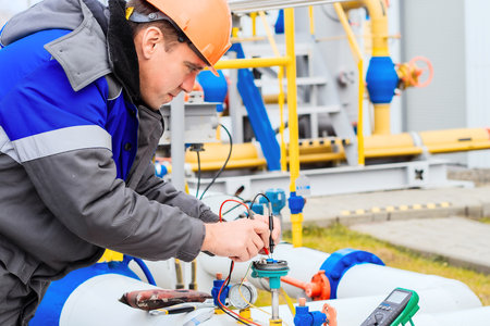 A Caucasian electrician in a helmet measures the voltage on the sensors of a gas distribution station with a multimeter outside. Authentic workflow. Gas pipeline maintenanceの写真素材
