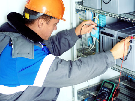 Professional electrician checks industrial equipment with multimeter. Worker in hard hat and winter coat measures battery charge of emergency substation and looks at readings of multimeter. Authentic workflow.の写真素材