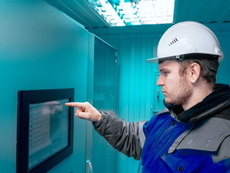 Engineer in hard hat and winter pea coat presses his finger on touch screen of modern production equipment indoors. Setting up automatic control system. Gas distribution station operator monitors gas flow readings. Photo. side view.の写真素材