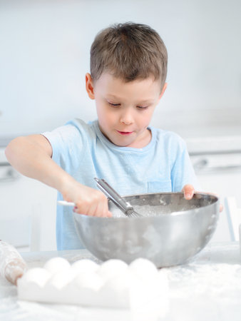 Caucasian boy sits at kitchen table and prepares dough from flour and eggs in large metal cup. Child plays cook in kitchen.の写真素材