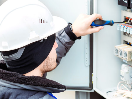 Professional electrician in hard hat connects wires with screwdriver in switchboard on winter day. Close-up portrait. Back view of head.の写真素材