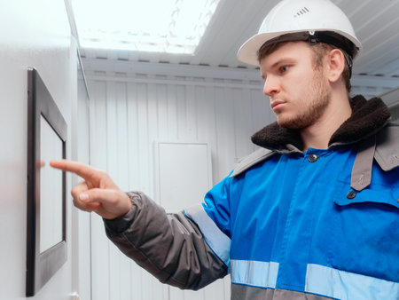 Engineer in hard hat and winter pea coat presses his finger on touch screen of modern production equipment indoors. Setting up automatic control system. Gas distribution station operator monitors gas flow readings. Photo. side view.の写真素材