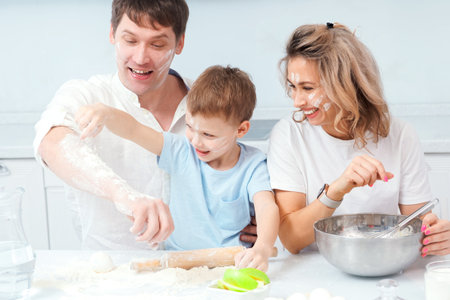 Young family with child prepares lunch together in home kitchen. Cheerful parents and son sit at table and get dirty in flour. Happy family at home. Style of life.の写真素材