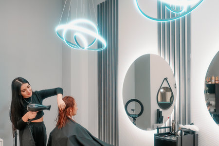 Woman hairdresser stylist doing hair styling by blow dryer to woman with red hair in modern beauty salon. Hairdressing salon.の写真素材