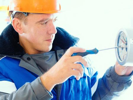 An electrician or instrumentation fitter with a screwdriver in his hand adjusts the operation of a pressure gauge at an indoor gas industrial facility. Caucasian worker in an orange hardhat and a winter pea coat at work. Close-up portrait.の写真素材