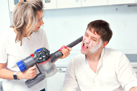 Young family couple have fun in home kitchen. Wife cleans her husbands face, which is covered in flour, with vacuum cleaner. Man covered his face in flour. Funny photo.の写真素材