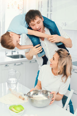 Young caucasian family mom dad and child are preparing birthday cake in home kitchen and having fun and fooling around. Cheerful parents together with their son start dough at kitchen table.の写真素材