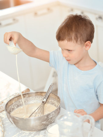 Cheerful boy plays chef at the table in the home kitchen. Caucasian child kneads the dough in a metal bowl. Playful kid in the kitchen.の写真素材