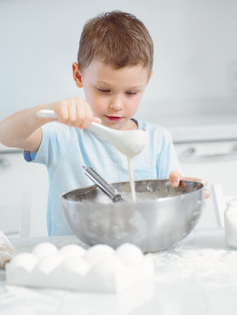 Caucasian boy sits at kitchen table and prepares dough from flour and eggs in large metal cup. Child plays cook in kitchen.の写真素材