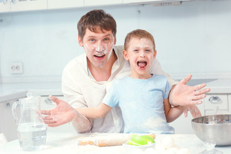 Father and son are preparing lunch or birthday cake together in home kitchen at table. Dad and child got dirty in flour and have fun in kitchen. Caucasian man with small child look at camera and have fun. Funny portrait of parent with little boy.の写真素材