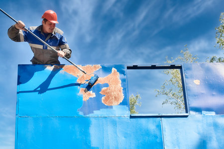 An industrial professional painter in a hard hat and work clothes paints a metal surface with a roller on a summer day.の写真素材