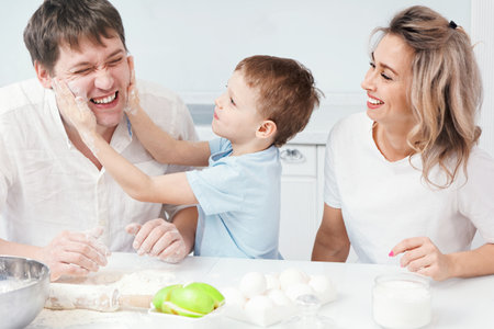 Portrait of a cheerful happy caucasian family at home in the kitchen. Mom dad and child cook dinner and have fun. The boy stained his fathers face with flour and laughs. Funny photo. Young family of threeの写真素材