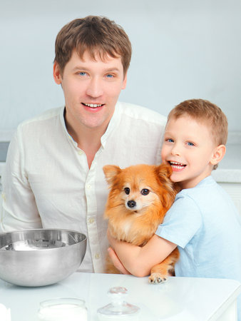 Young dad with small son and dog are sitting at table in kitchen, looking at camera and smiling.の写真素材