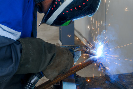 Masked welder works in workshop and sparks fly. Close-up portrait. Industrial background. Authentic workflow. Welder welds pipe.の写真素材