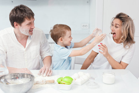 Child and parents are preparing pie from dough in kitchen and got dirty in flour. Young caucasian family fooling around and having fun at home cooking table. Cheerful photo.の写真素材