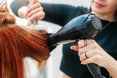 Hair styling with hairdryer in beauty salon. Close-up. Stylist hairdresser combs red hair of client and does hairstyle. Hands with hair dryer and comb.の写真素材
