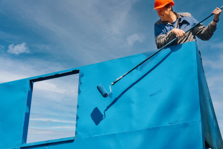 a professional painter in hard hat and work clothes paints metal structure with paint roller on summer day. Industrial painter, high up against sky, paints surface of wall. Real worker. Authentic workflow.の写真素材